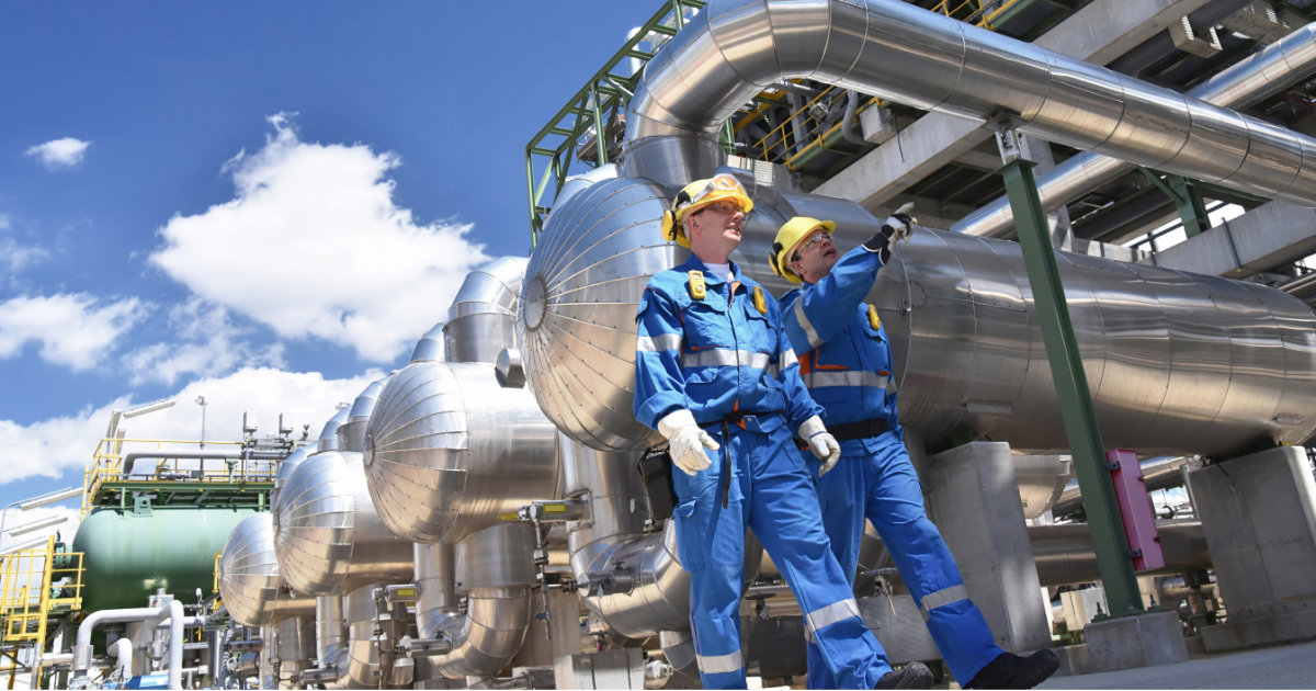 Two industrial workers in blue protective gear and yellow hard hats inspect piping at a large processing facility.